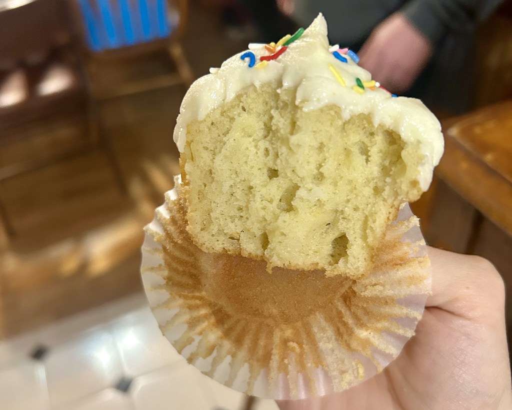A partially eaten cupcake with creamy frosting and colorful sprinkles, held in a hand. The cupcake wrapper is visible, and the background includes blurred wooden furniture.