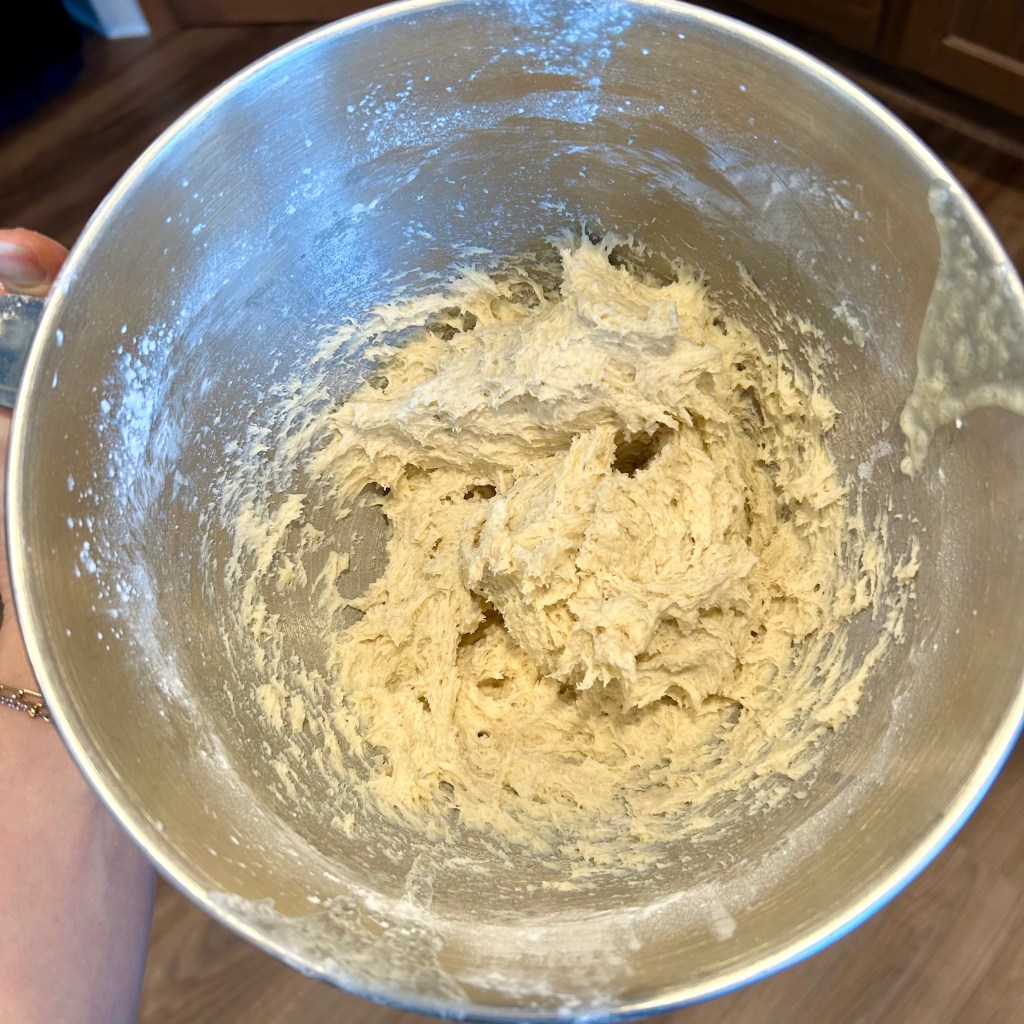 A mixing bowl filled with cookie dough, featuring a smooth and creamy texture, with a hand holding the bowl. The background shows a wooden countertop.