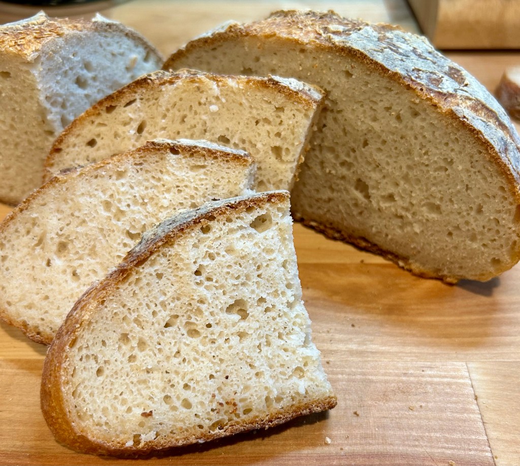 Sliced artisanal bread with a golden crust and soft inside, arranged on a wooden cutting board.