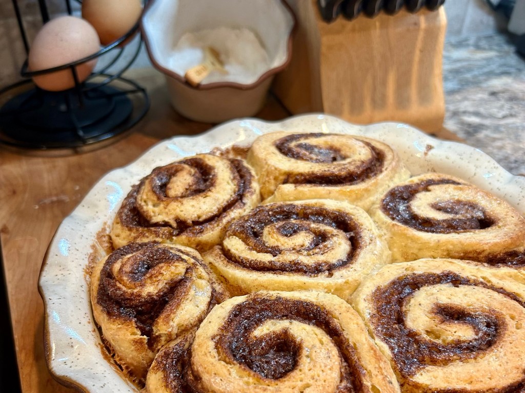 Freshly baked cinnamon rolls arranged in a circular dish, with a rustic kitchen background featuring eggs and sugar.