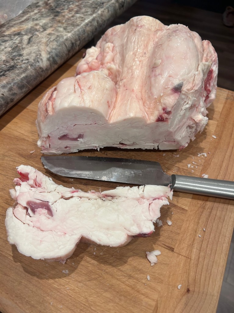 A chunk of raw fatback on a wooden cutting board with a stainless steel knife next to it, showing a cut piece of the fat.