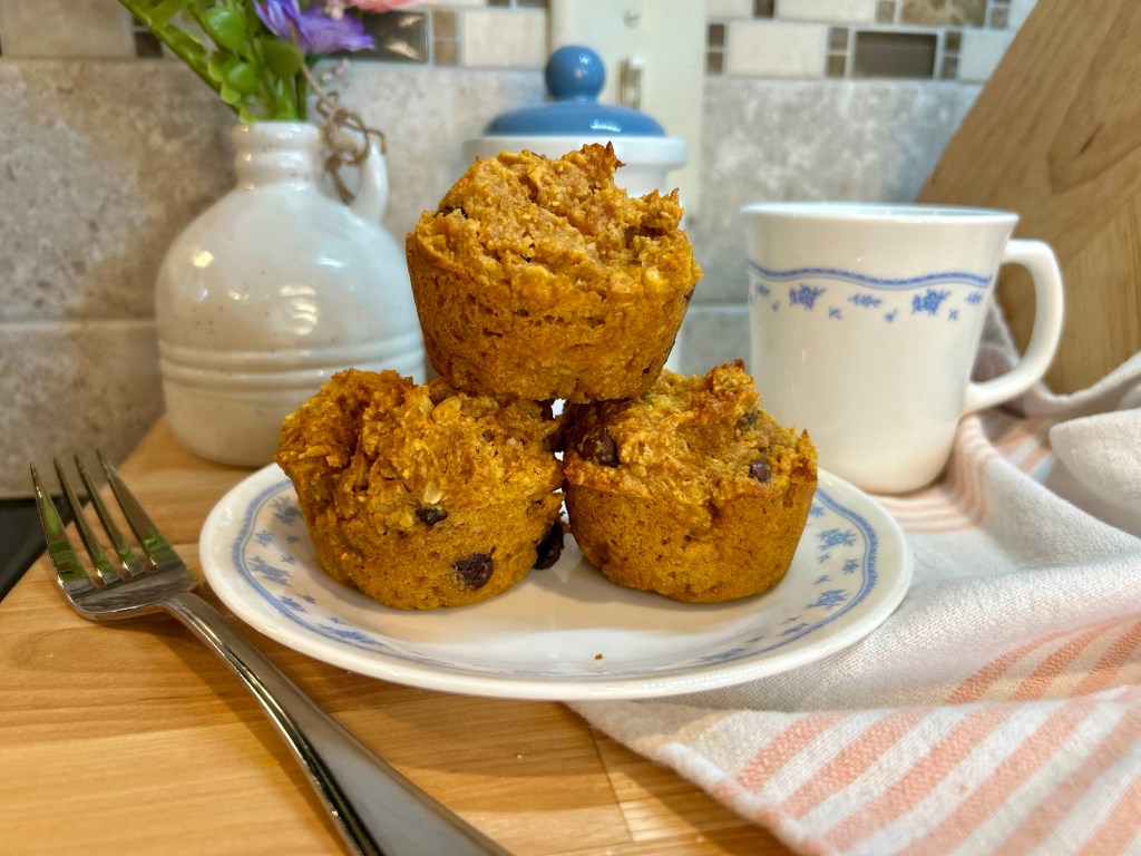 Three freshly baked muffins stacked on a blue and white patterned plate, accompanied by a fork and a white ceramic mug, with a vase of flowers in the background.