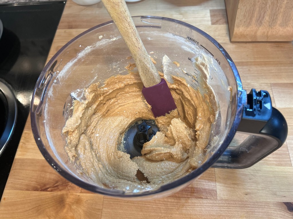 A food processor bowl containing a thick, creamy batter with a spatula resting inside, set on a wooden countertop.