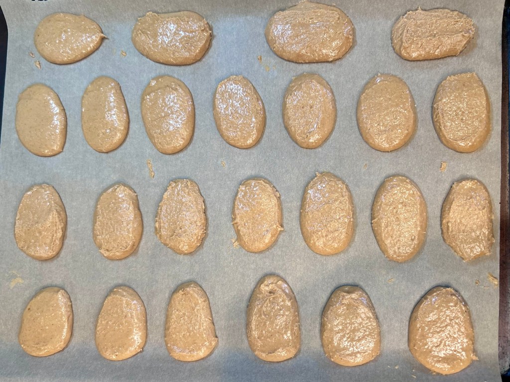 Baking sheet lined with parchment paper, covered with evenly shaped dollops of a tan-colored batter.