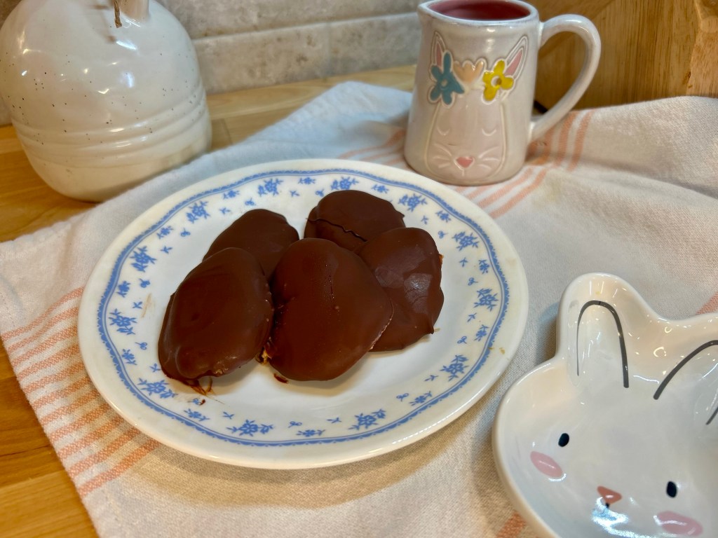 A plate of chocolate-covered snacks sits on a cloth, with a bunny-themed dish nearby and a mug decorated with a rabbit design in the background.