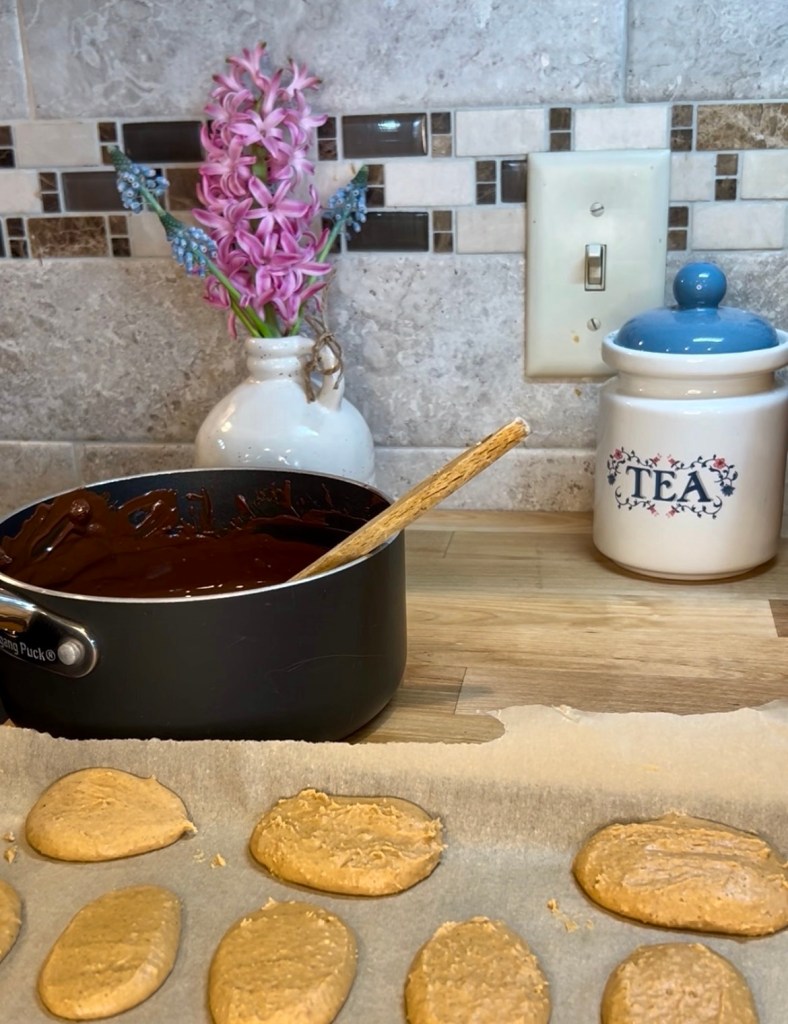 A kitchen countertop with a pot of melted chocolate, a wooden spoon, and freshly shaped cookies on parchment paper. A vase with pink flowers and a decorative tea container are also visible in the background.