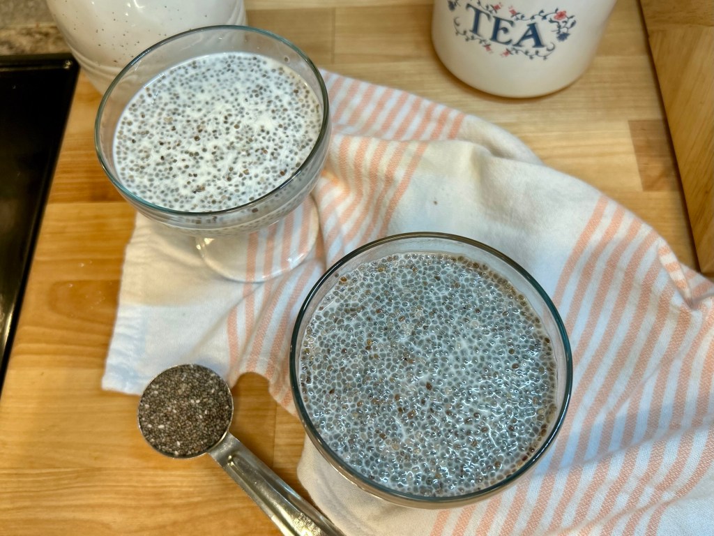 Two glass bowls of chia pudding on a wooden countertop, accompanied by a measuring spoon filled with chia seeds and a striped cloth.