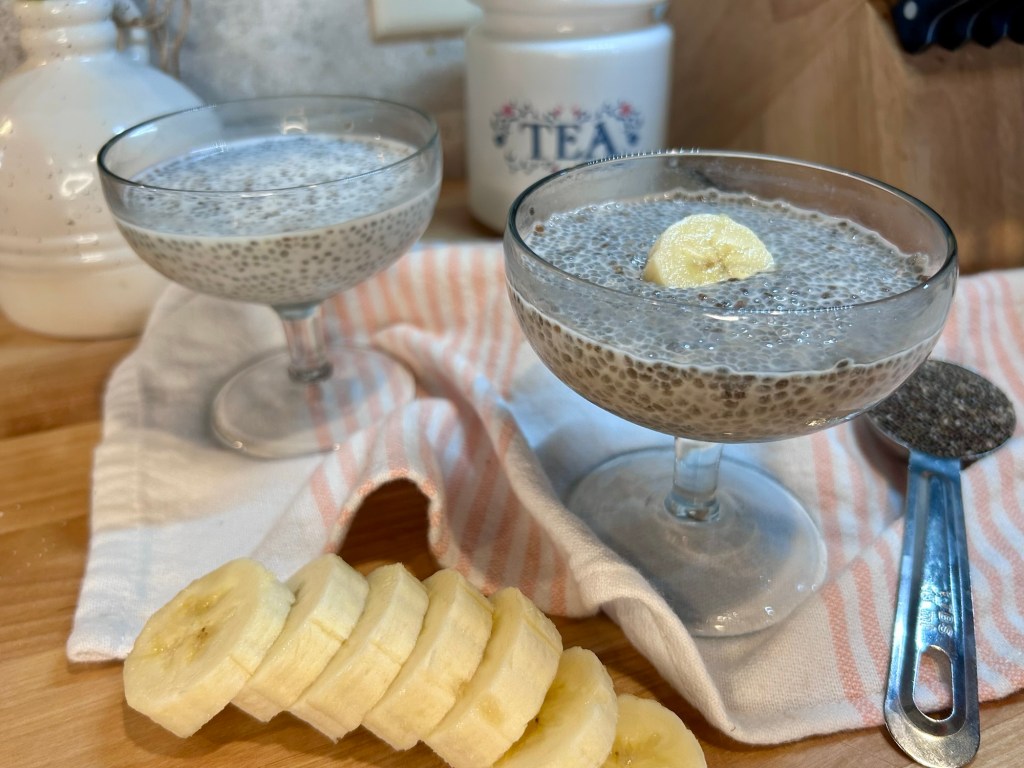 Two glasses of chia seed pudding topped with banana slices, placed on a wooden surface with a striped napkin and a spoon of chia seeds in the background.