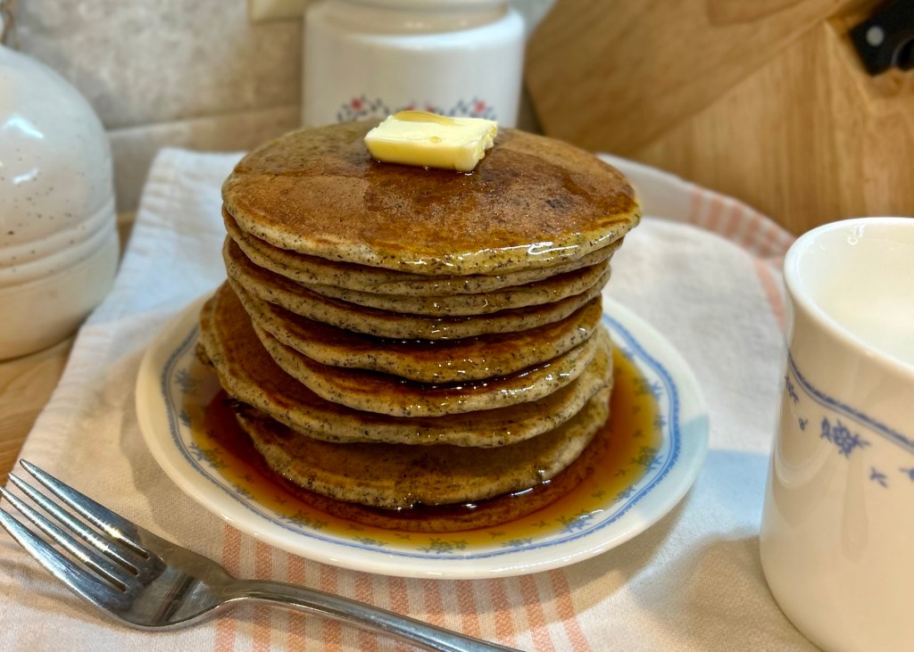 A stack of golden-brown pancakes topped with a pat of butter and drizzled with syrup on a white plate, accompanied by a fork and a cup.