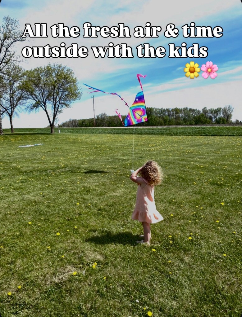A child with curly hair wearing a pink dress stands in a grassy field, flying a colorful kite on a sunny day.