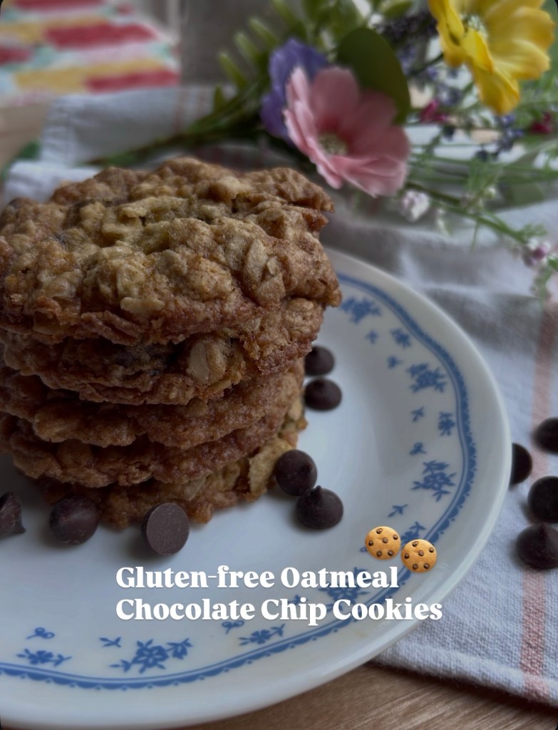 A stack of gluten-free oatmeal chocolate chip cookies on a decorative plate, accompanied by chocolate chips and surrounded by a floral arrangement.