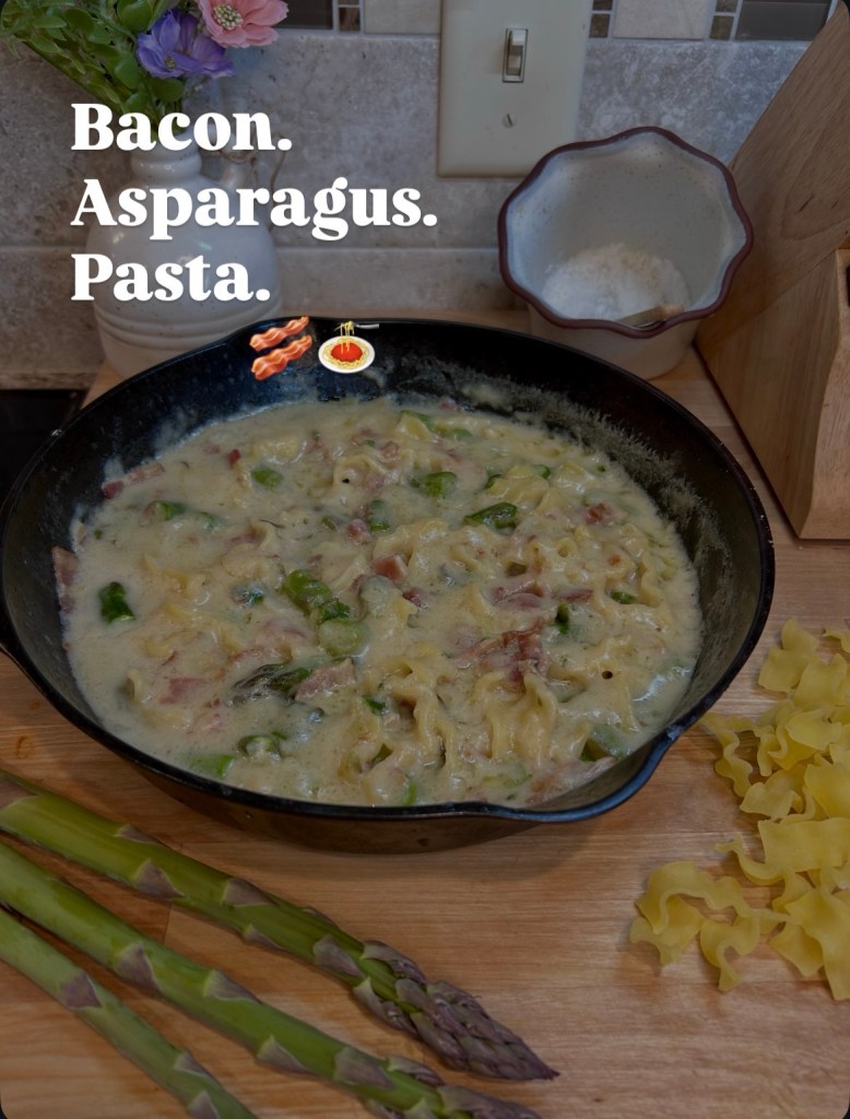 A creamy pasta dish with bacon and asparagus in a skillet, alongside fresh asparagus and uncooked pasta on a wooden surface.
