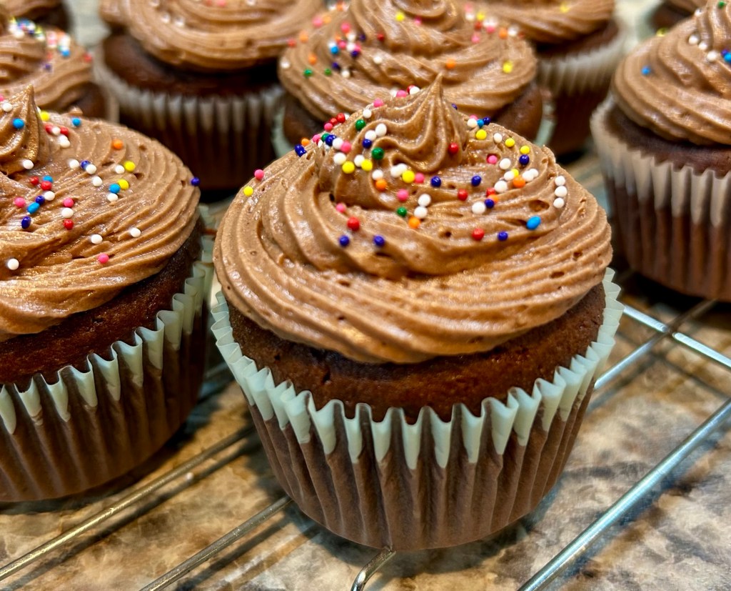 Close-up of chocolate frosted cupcakes decorated with colorful sprinkles, arranged on a cooling rack.