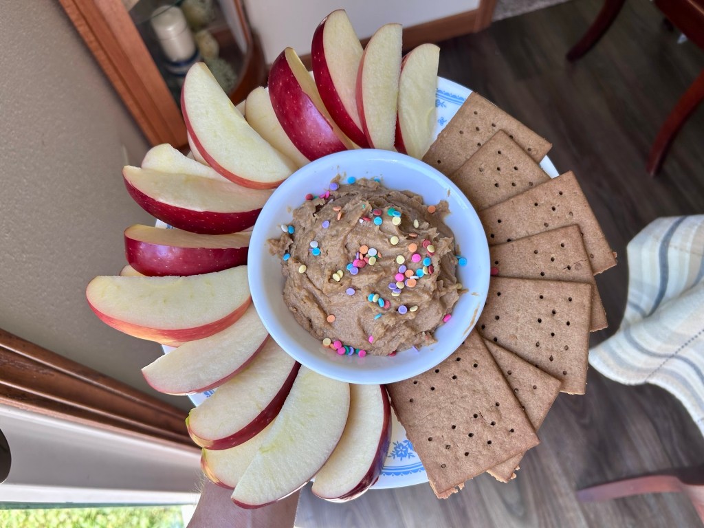A plate arranged with apple slices, a bowl of dip topped with colorful sprinkles, and graham crackers.